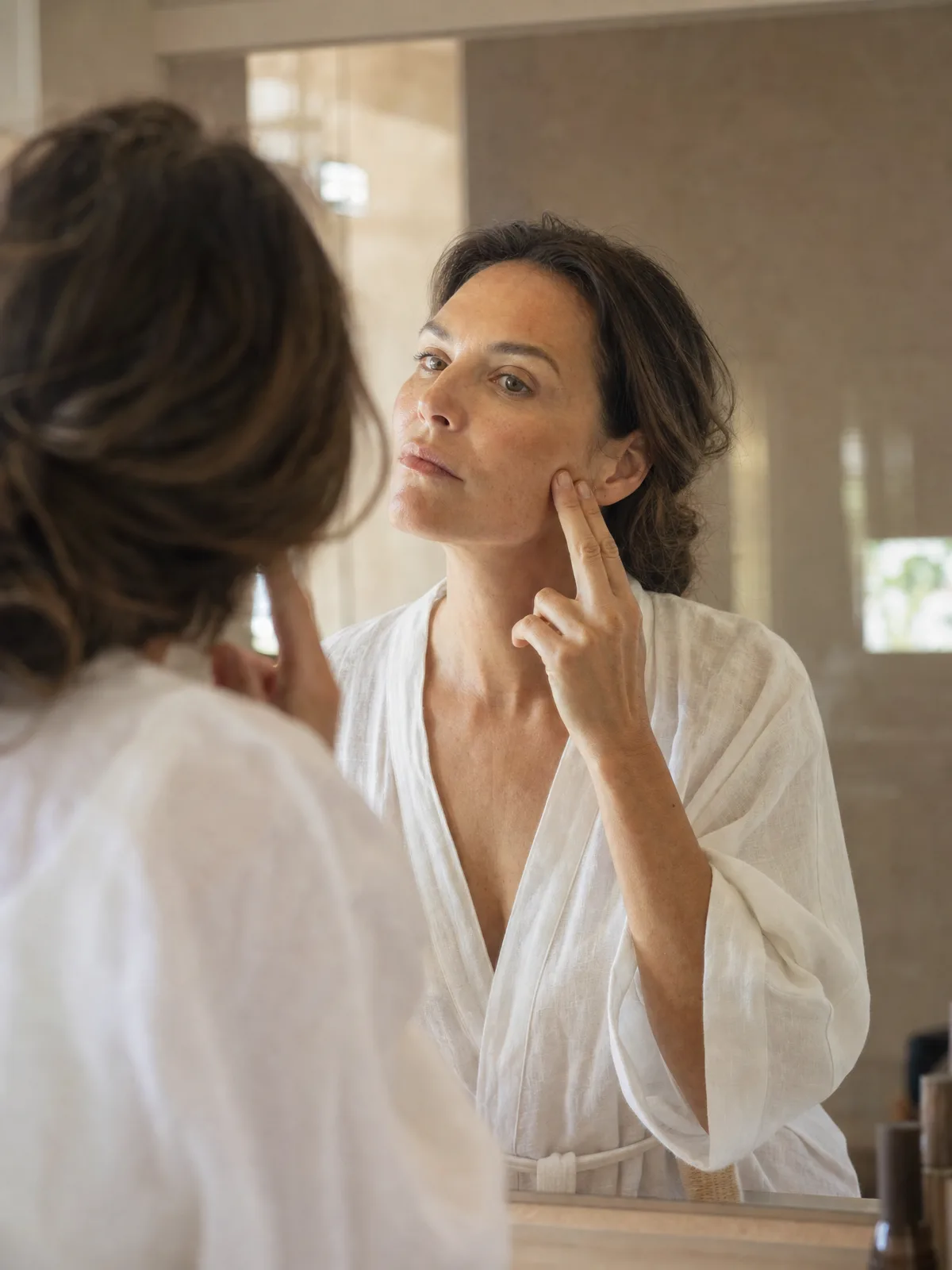 Woman in her forties at her bathroom mirror, two fingers on her cheek, studying her jawline, in Laval