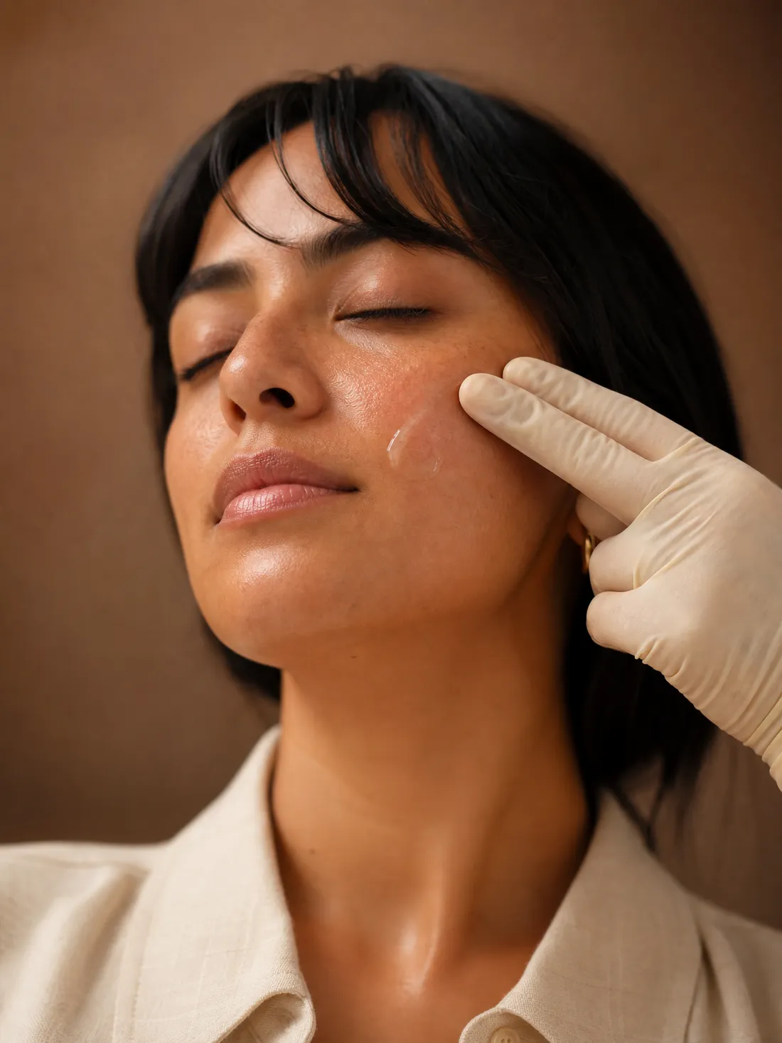 A gloved hand touches the cheek of a woman with her eyes closed, right after a microneedling session